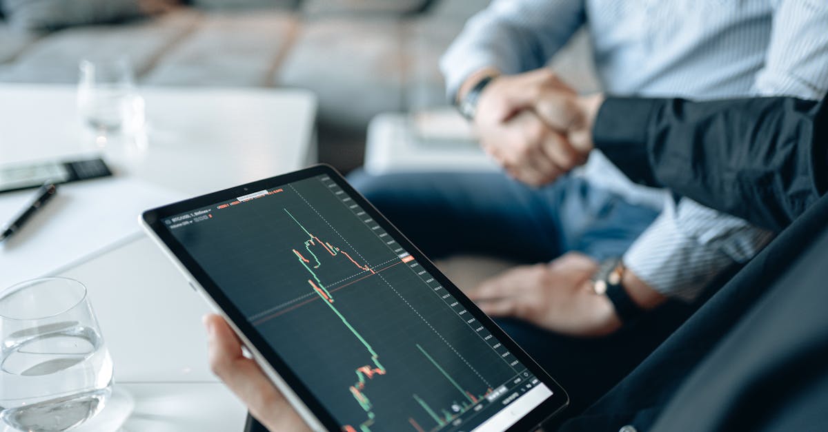 Close-up of tablet showing stock graph during business meeting with handshake in background.